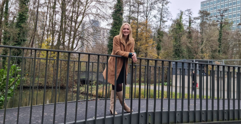 Een vrouw staat leunend op de leuning van een metalen voetgangersbrug in een parkachtige omgeving. Ze draagt een lange camel­kleurige jas en laarzen. Op de achtergrond zijn bomen met herfstkleuren en enkele hoge kantoorgebouwen te zien.