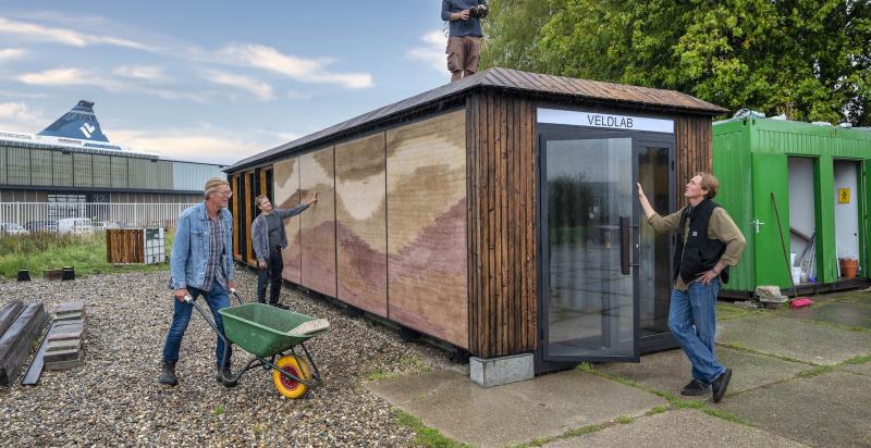 Drie mensen werken rond het Veldlab; een persoon staat op het dak en twee anderen bekijken de buitenzijde van de met natuurlijke materialen geïsoleerde zeecontainer.