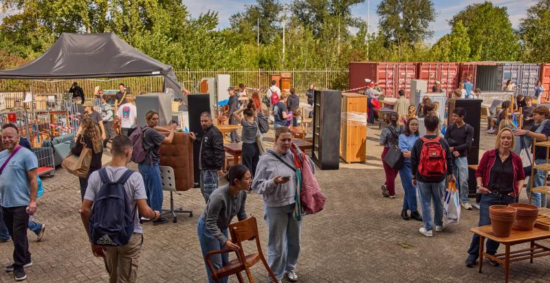 Druk buitenterrein waar bezoekers tussen uitgestalde tweedehands meubels en woonspullen rondkijken.