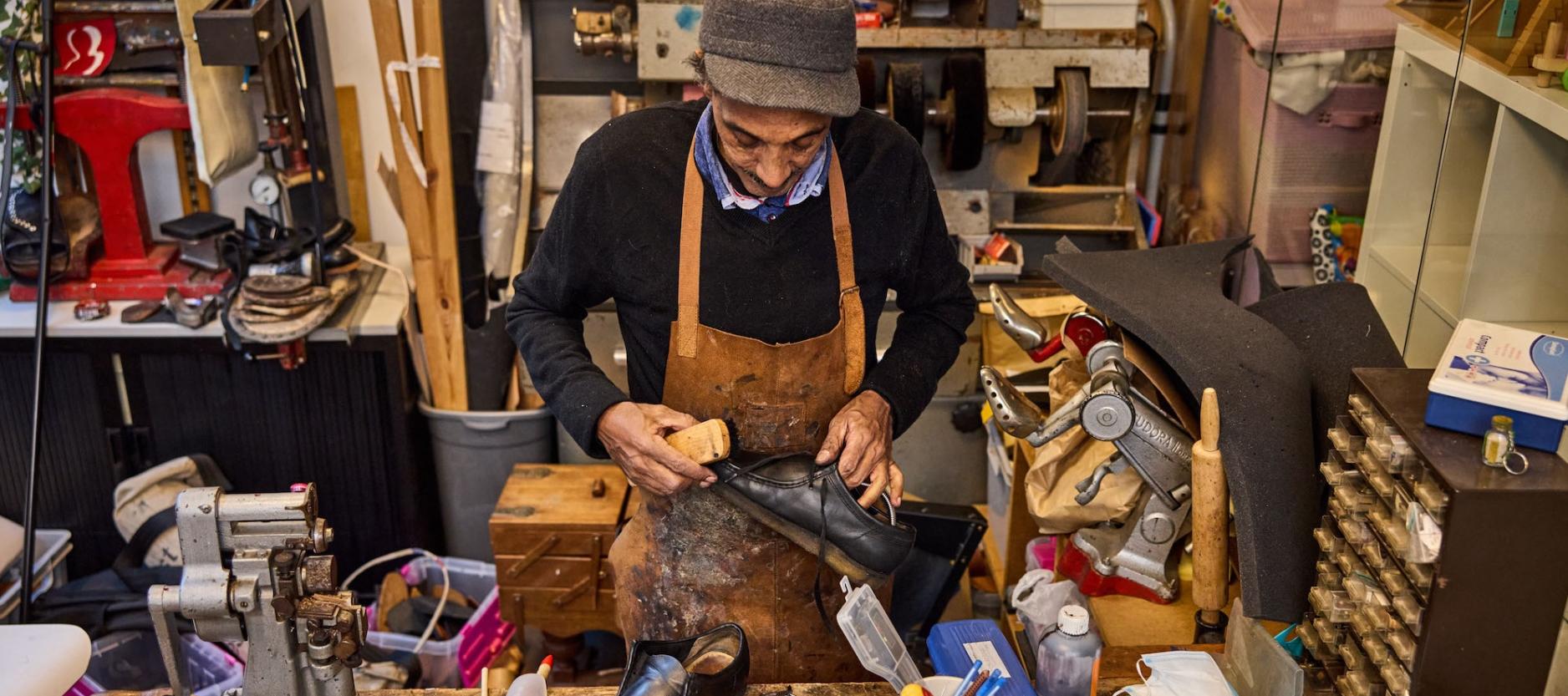 Benyamin Tesfai aan het werk in zijn werkplaats. Hij draagt een leren short en pet. De houten werkbank ligt vol met gereedschap.