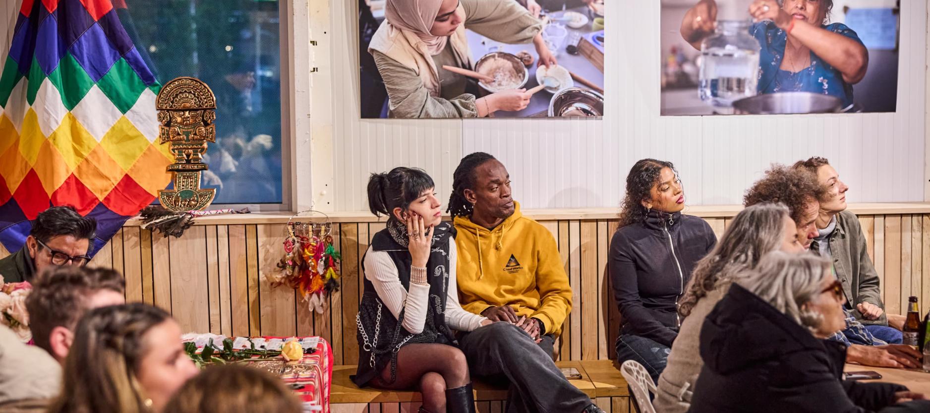 Groepje mensen zit in de tentoonstellingsruimte in De HER te luisteren naar een spreker op het podium.