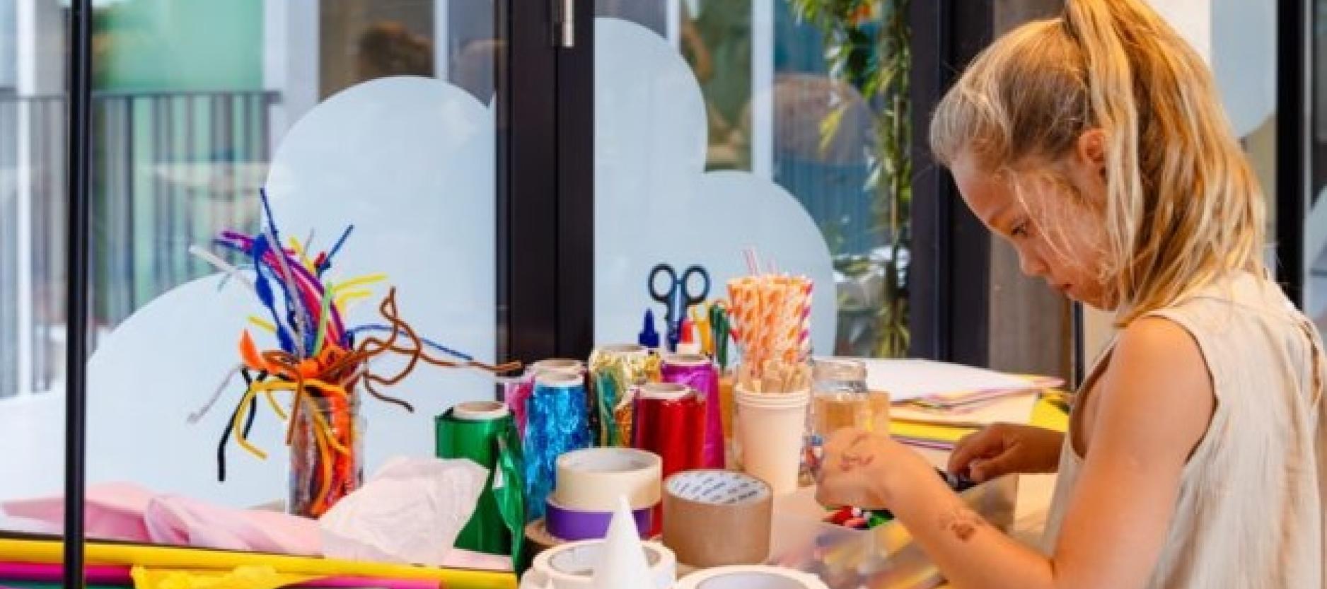 Een meisje staat aan een tafel vol met kleurrijke knutselmaterialen