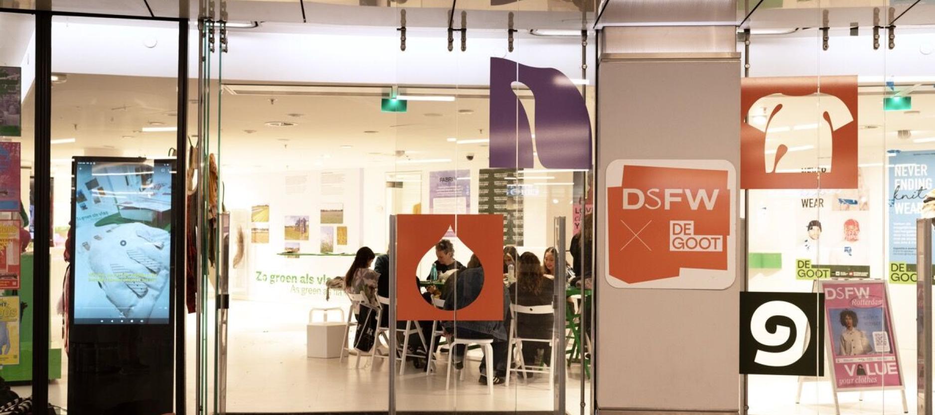 ezoekers zitten aan tafels in een lichte winkelruimte tijdens een evenement van DSFW, met kleurrijke campagne-uitingen bij de ingang.