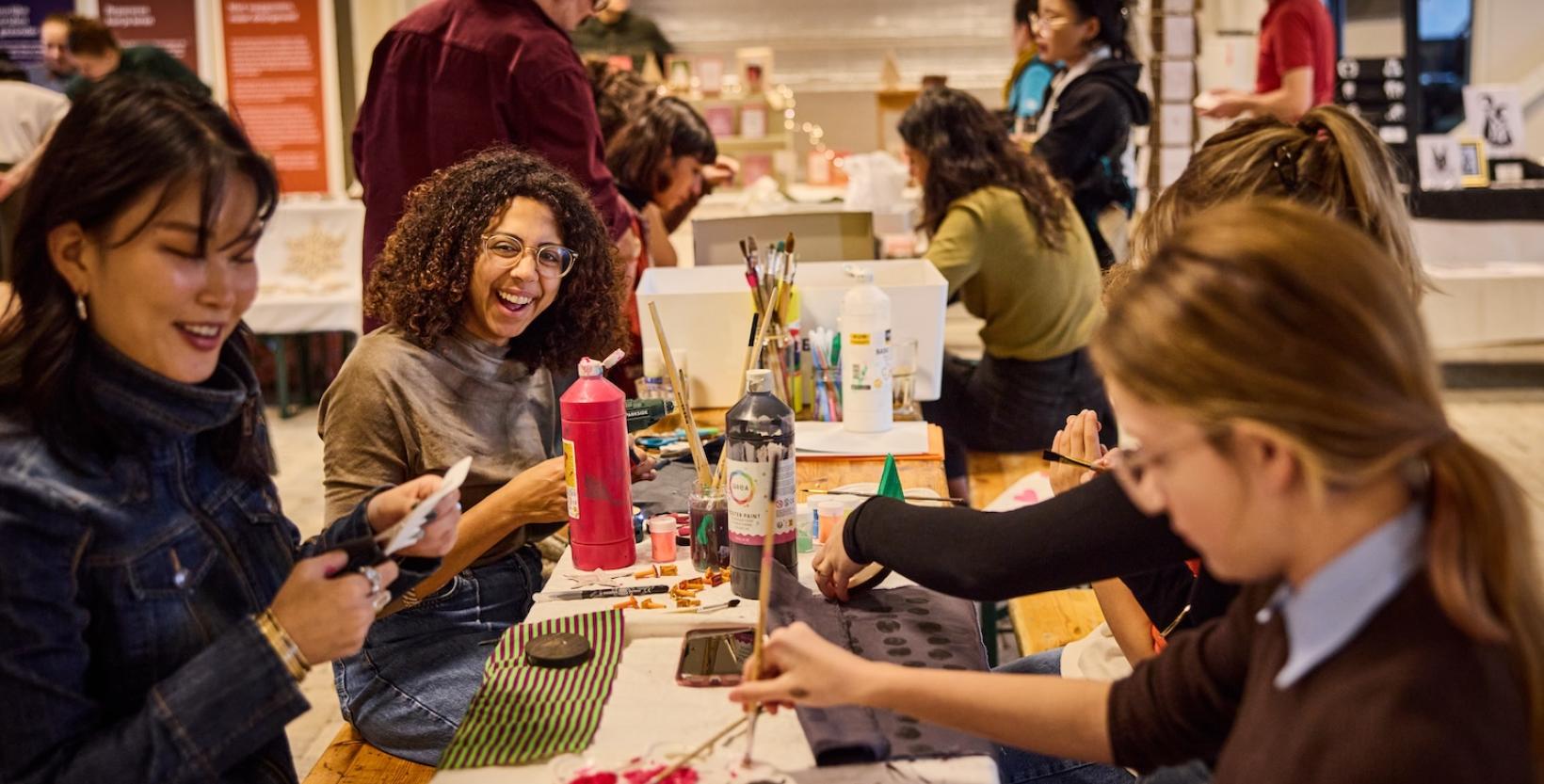 Bezoekers nemen deel aan een creatieve workshop en beschilderen textiel en objecten aan een grote werktafel.